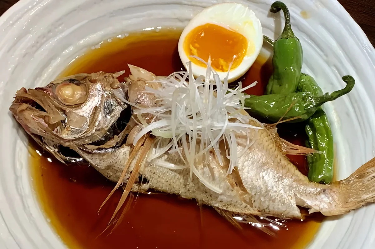 A whole simmered Japanese fish (Nizakana) on a white plate. It is topped with finely shredded white leek and served with a soft-boiled egg and green shishito peppers.